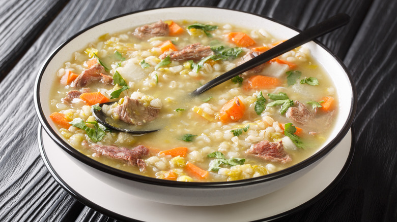 bowl of scotch broth with meat, barley, vegetables, with a spoon against dark background