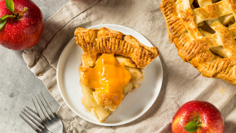 Slice of apple pie plated with melted cheddar cheese on top, on a table with whole apples