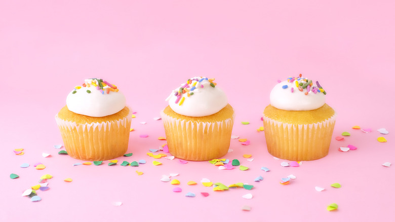 Three yellow cupcakes with white frosting and sprinkles on a bright pink background