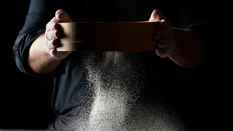 Person sifting flour using a fine mesh sieve on a black background