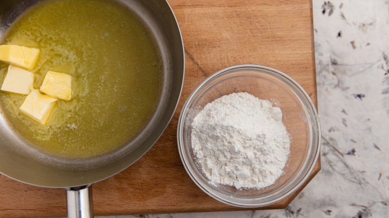Melted butter in a pan and flour in a bowl on a wooden board to prepare a roux