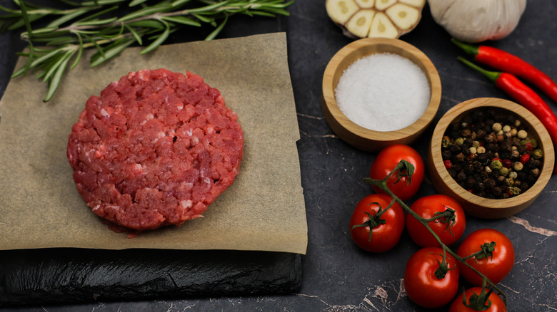 Raw ground beef patty, with salt, pepper, and vine tomatoes sitting next to it