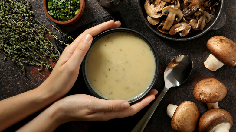 A bowl of cream of mushroom soup is cupped between two hands, with ingredients surrounding it