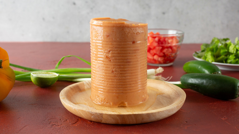 A block of refried beans from a can, still holding the shape of the can itself, sits in a wooden bowl surrounded by vegetables