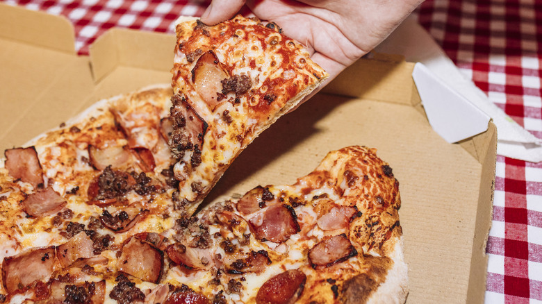 A person lifts a slice of meat pizza from a takeout box