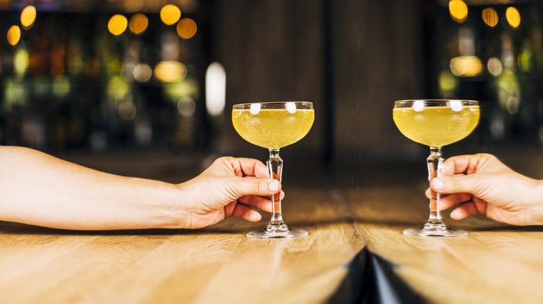 A hand holds a cocktail in a coupe glass, with a mirror reflecting it