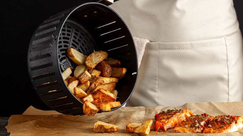 A chef tips potatoes out of an air fryer onto parchment paper, next to some cooked salmon fillets
