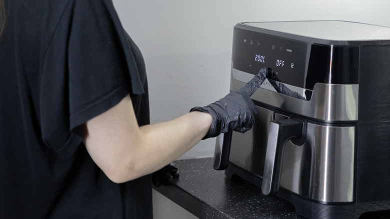 A person wearing a black rubber glove taps an air fryer's front panel to change its temperature settings