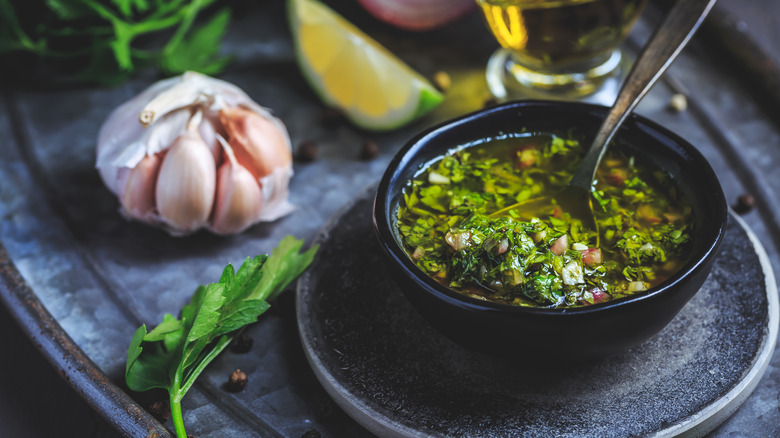 Serving dish of chimichurri surrounded by herbs, garlic, lemon, and oil