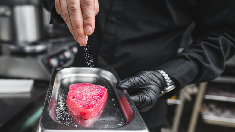 A chef sprinkles salt onto a tuna steak in a kitchen