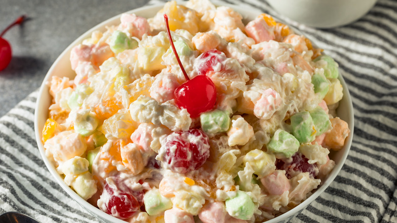 a bright bowl of ambrosia salad garnished with a maraschino cherry on top of a striped towel.