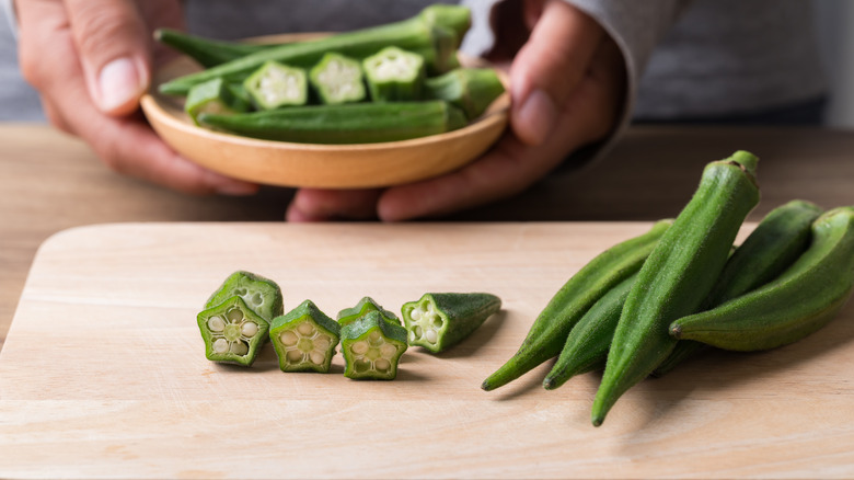 a board with pods of okra and slices of okra. In the background, a person's hands hold a bowl of okra pods and sliced okra.