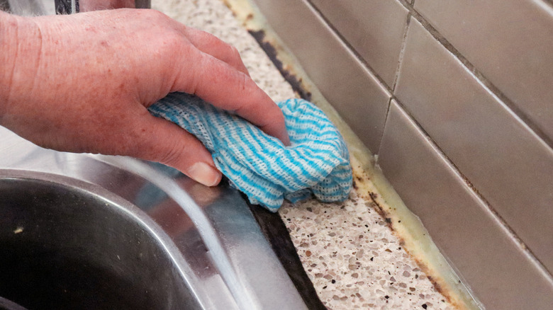 A person cleans mold from behind a kitchen sink