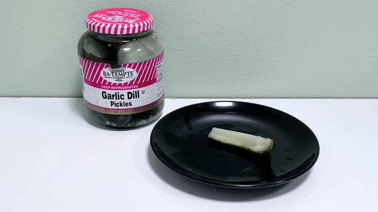 A dill pickle on a black plate next to a jar of Ba-Tampte Garlic Dill Pickles