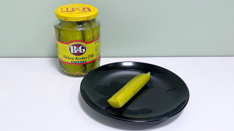 A dill pickle on a black plate next to a jar of B&G Deluxe Kosher Dill