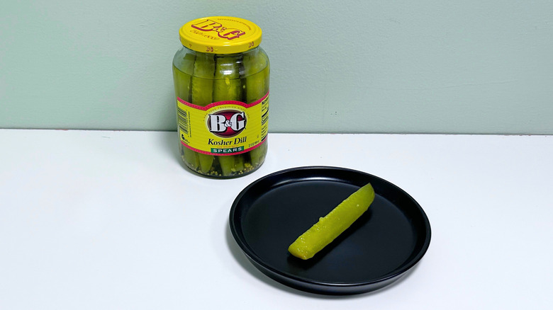 A dill pickle on a black plate next to a jar of B&G Kosher Dill Spears