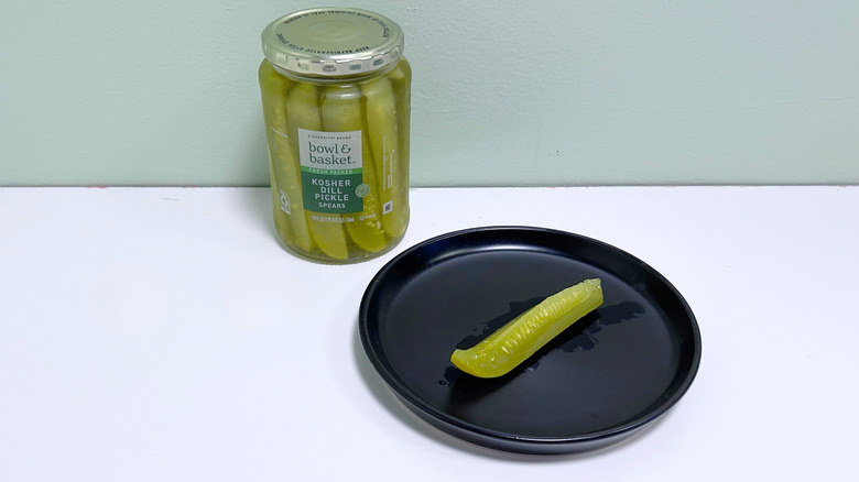 A dill pickle on a black plate next to a jar of Bowl and Basket Kosher Dill Pickle Spears