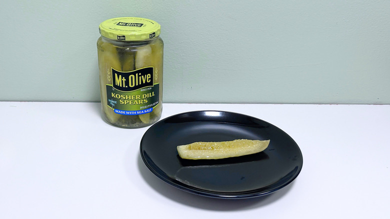 A dill pickle on a black plate next to a jar of Mt. Olive Kosher Dill Spears