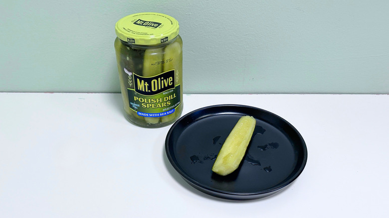 A dill pickle on a black plate next to a jar of Mt Olive Polish Dill Spears