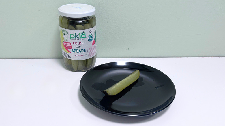 A dill pickle on a black plate next to a jar of pkld Polish Dill Spears