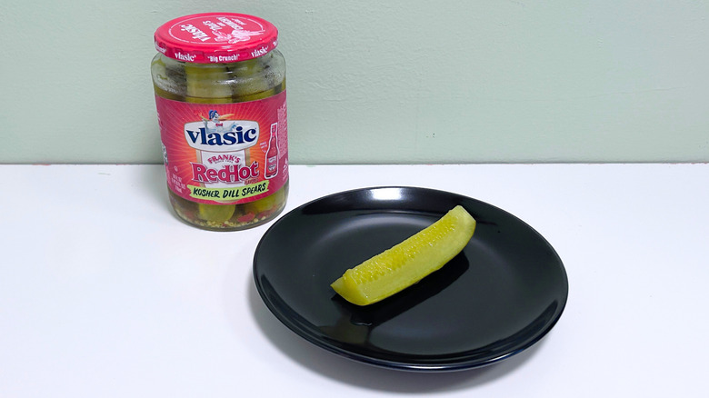 A dill pickle on a black plate next to a jar of Vlasic Frank's Red Hot Kosher Dill Spears