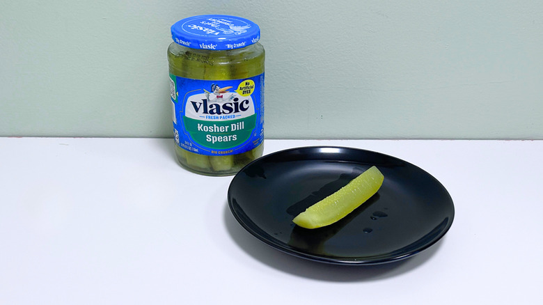 A dill pickle on a black plate next to a jar of Vlasic Kosher Dill Spears