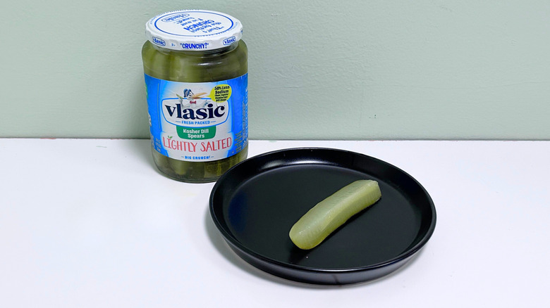 A dill pickle on a black plate next to a jar of Vlasic Lightly Salted Kosher Dill Spears