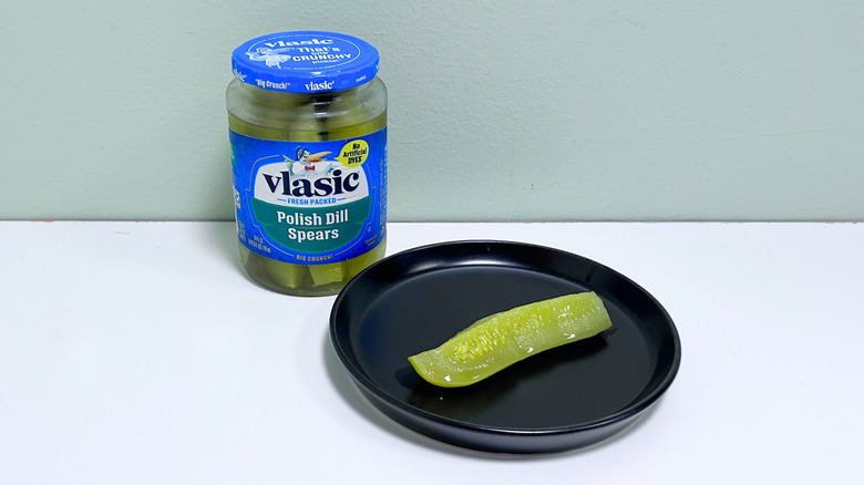 A dill pickle on a black plate next to a jar of Vlasic Polish Dill Spears