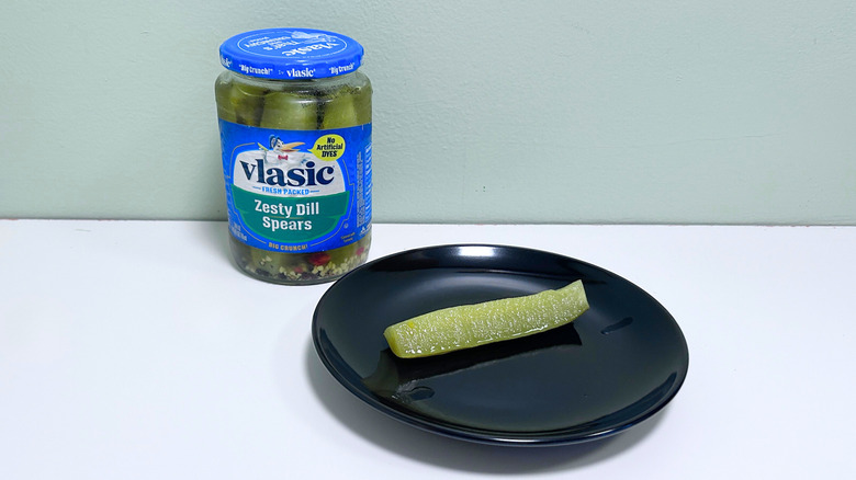 A dill pickle on a black plate next to a jar of Vlasic Zesty Dill Spears