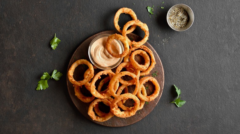 A stack of onion rings on a wooden board, with dipping sauce and spice mix
