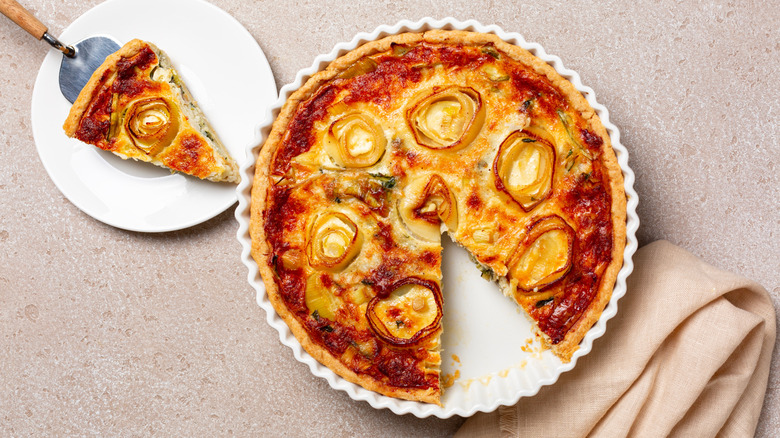 Homemade onion quiche, with a slice cut from it sitting on a white plate