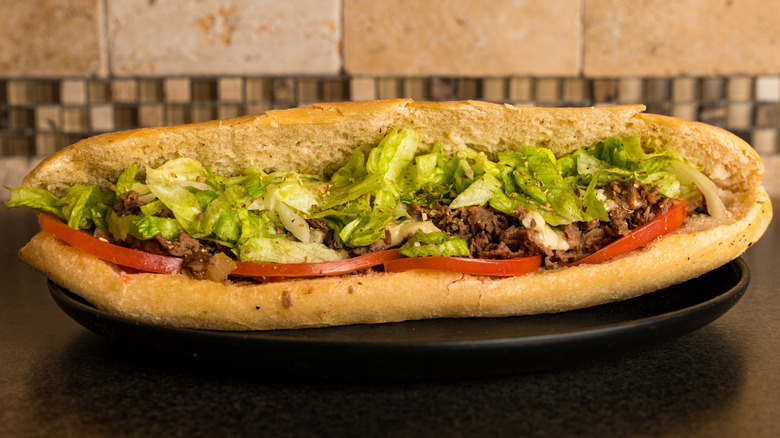 Cheesesteak hoagie with lettuce and sliced tomato, on a black plate