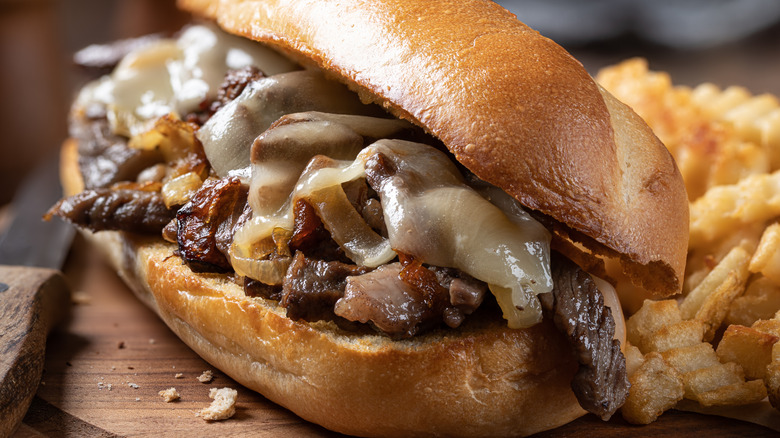 Close up of a Philly cheesesteak on a toasted roll with caramelized onions and a side of crinkle-cut fries