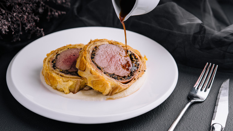 Slices of beef Wellington on white plate with red wine sauce being poured on top