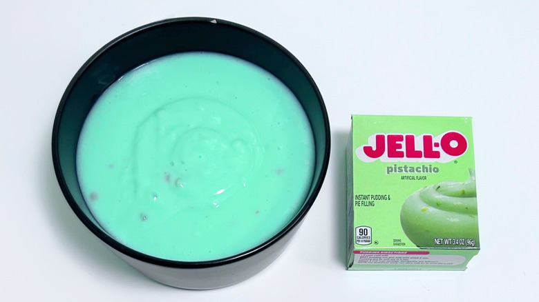 A bowl of bright teal pudding next to a box of Jell-O Pistachio Pudding Mix
