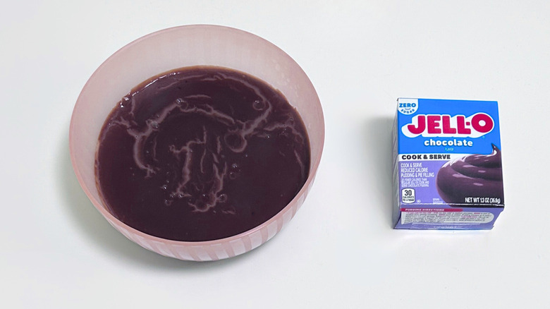A bowl of pudding with a dark brown skin on top, next to a box of Jell-O Zero Sugar Cook and Serve Chocolate Pudding Mix