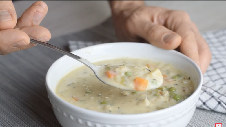 a person spoons a bite of oatmeal stew with carrots and peas out of a white bowl