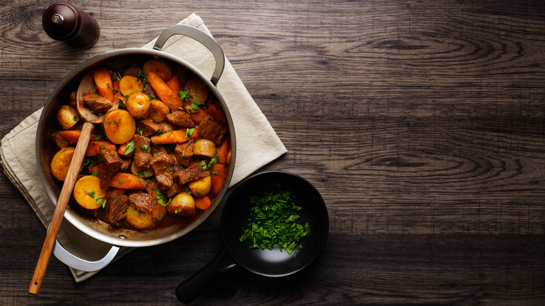 a pot of braised beef with potatoes and carrots with a side bowl of chopped parsley and a pepper grinder