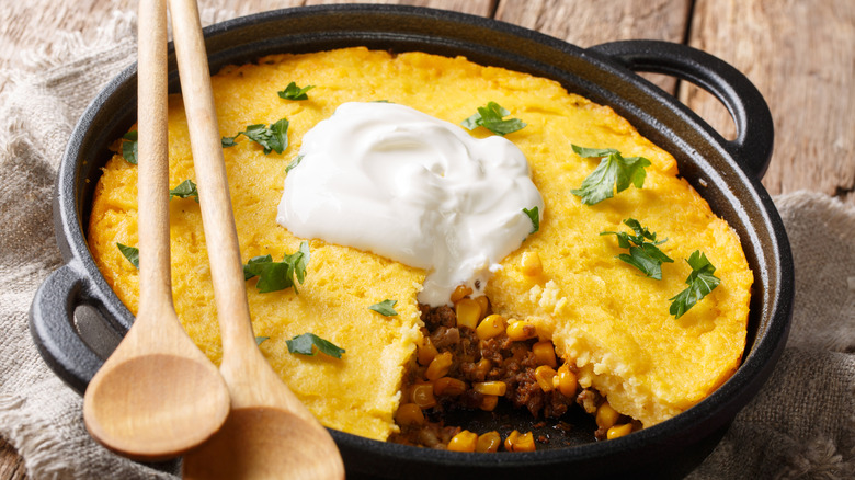 a cast iron bowl full of tamale pie with one slice missing with a filling of ground beef and corn and garnished with cilantro and sour cream
