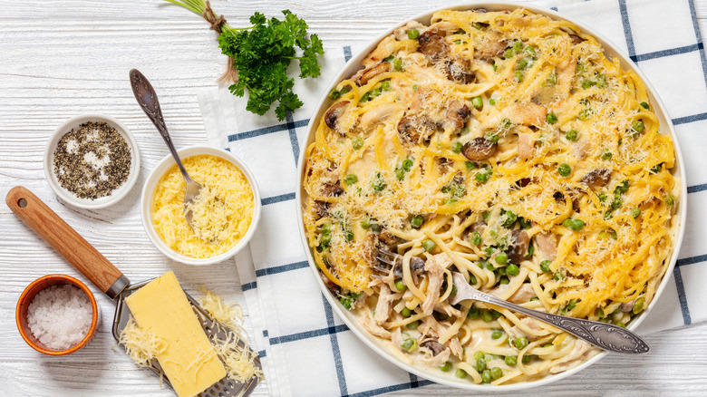 a large bowl of tetrazzini on a white table with a blue checked napkin and sides of salt, pepper, grated Parmesan, and parsley