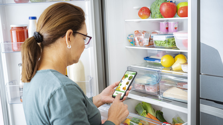 woman scrolls through grocery store app while looking in fridge