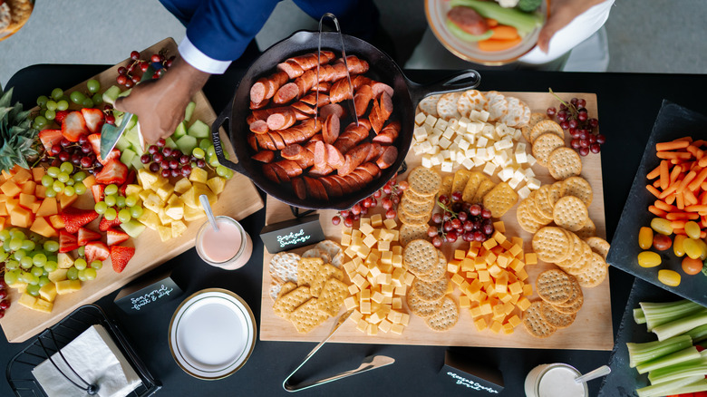 A buffet table with mixed fruit, cheese and crackers, and sliced smoked sausage.