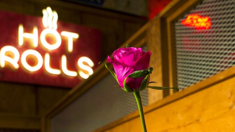 A lit-up Hot Rolls sign at Logan's Roadhouse with a pink rose in the foreground.