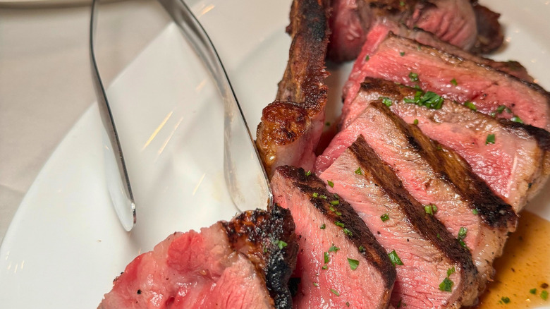 A carved steak on a white plate with serving tongs and amber-colored pan sauce.