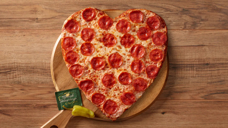 A Papa John's heart-shaped pepperoni pizza on a wooden board with a banana pepper and seasoning packet.