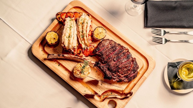 Two lobster tails and a bone-in steak on a wooden board.