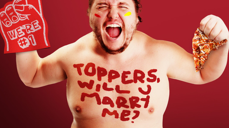 A man with red and yellow athletic face paint and a foam finger holding a pizza slice the words "Toppers, will u marry me?" written on his chest.