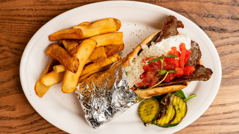 Gyro, steakfries, and pickles on a white plate against a wood background