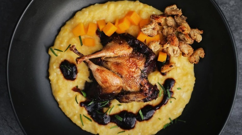 A black bowl of sofkee topped with diced carrots and a whole roast quail
