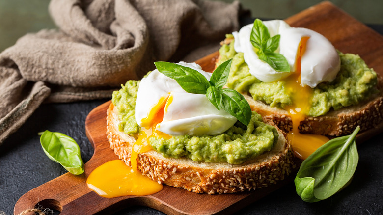 Two slices of avocado toast topped with poached eggs on a wooden cutting board